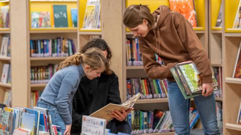 A young girl and a woman look at books in a library. They are surrounded by shelves holding colourful books.