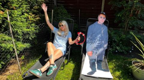 Liv Bridge reclines on a garden chair while holding a tambourine in her arm. Next to her is a cardboard cut out of Liam Gallagher. Liv is blonde, and is wearing a Oasis band t-shirt and dark sunglasses.
