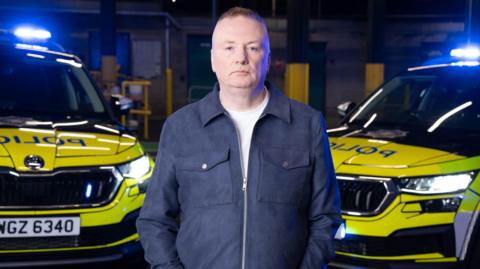 Stephen Nolan looking at the camera, he has brown hair. He is wearing a navy jacket that is zipped up halfway with a white t-shirt under it. Behind him are two police cars that have blue lights on.