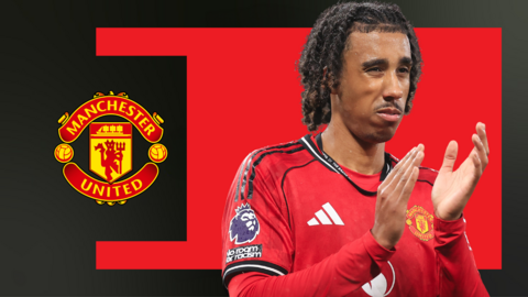 Leny Yoro, wearing Manchester United home kit and applauding towards fans, in front of a red and black background and the club badge