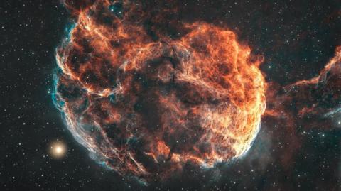 The Jellyfish Nebula is a circular ball of brightly coloured gases. It has elements of red, blue, black and orange and is set in a night sky spotted with stars. 