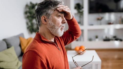 Worried man with painful facial expression sitting on living room couch and holding hand on his forehead. He is wearing an orange top. 