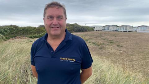 A man with light-brown hair is standing on long grass in front of a row of cream-coloured caravans with decking. The clouds behind him are grey. He is wearing a navy polo shirt which reads 'Coast and Country Holiday Parks.' He has his arms behind his back. The land behind him looks dry and has a few yellow flowers on it.