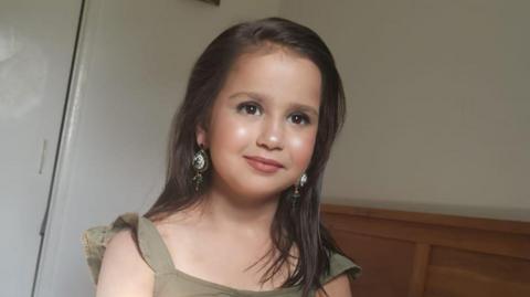 A young girl with straight brown hair and a green dress. She is smiling and has big sparkly earrings. 