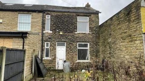 A dilapidated looking front of a house with lots of weeds growing in the garden, a tall brick wall to one side, and a fence to the other.