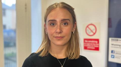 Aimee is a young woman with shoulder length blonde hair. She is wearing a black t-shirt, a silver necklace and silver earrings and is looking into the camera. She is stood in front of a door and white wall with a no-smoking sign on it out of focus behind her.