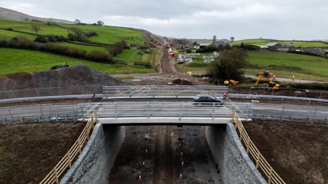 The new bridge with a car in motion travelling on it. The bridge is over an unfinished stretch of road, which looks like a dirt track, with machinery and workers in high viz working on the site.
