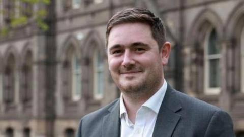 Chris Cooke in grey suit and white shirt with top button undone, smiling in front of an ornate building.