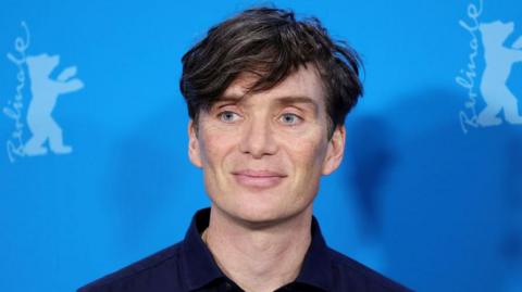 Cillian Murphy wearing a navy shirt smiling against a blue backdrop 