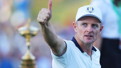 Ryder Cup European golfer Justin Rose wearing a white short sleeve top and a Ryder Cup hat. His right arm is pointing towards the camera with his thumb up. The Ryder Cup trophy is in silhouette in the background.