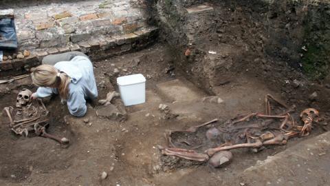 Skeletal remains lying in the soil bordered by a brick wall. A woman is on the ground next to one of the skeletons and there is a white bucket at her side. The woman has long blonde hair tied in a ponytail and is wearing a blue jumper and grey trousers. 