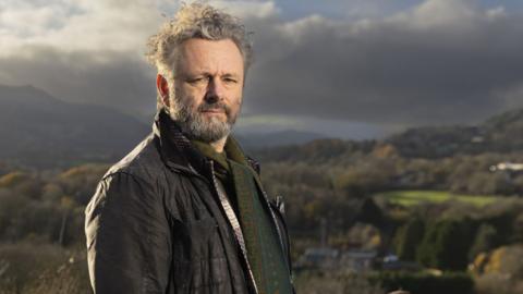 Standing on high ground above a valley, Michael Sheen looks towards the camera with rolling hills and patchwork fields stretching into the distance. He is wearing a brown jacket with a green patterned jumper. Sheen has grey hair and facial hair. Strong afternoon sunlight and heavy clouds create dramatic contrasts across the rural landscape.