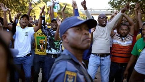 A police officer looks on as protesters chant slogans during protests in Mahikeng, in the North West province, on 20 April 2018