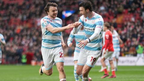 Ollie Rathbone, wearing a sky blue and white hooped Wrexham shirt, is congratulated by teammate Kieffer Moore after scoring against Charlton