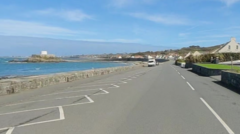 A picture of Route de la Lague and Route de Rocquaine. To the left of the road is the sea and Fort Grey. To the right are boundary walls, lawns and houses.