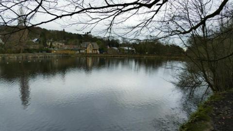 A large patch of water with overhanging trees and vegetation, At the far end of the water there are a collection of buildings.
