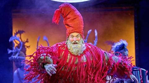 A man with a beard and a big red hat and costume on a stage with actors behind him 