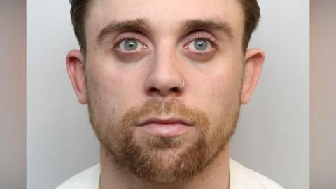 A police mugshot of Joe Martin who is staring blankly straight ahead. He has a short ginger beard and darker-coloured hair and a silver ring through his nose. His eyes are light blue. The background is pale grey and his white T-shirt is just visible. 
