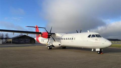 A white ATR 72-600 aircraft stationary at the airport. The aircraft is white with red at the back and on the wing. There is a building in the background and there are clouds in the sky.