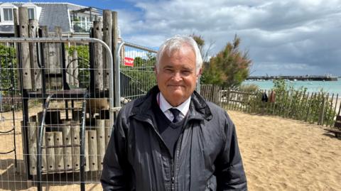 A person wearing a dark jacket, sweater, shirt, and tie stands on a sandy beach beside a fenced wooden playground structure. Behind the playground are seaside houses, while a turquoise sea, harbour wall, and cloudy sky are visible in the background. Bright sunlight illuminates the scene despite the approaching dark clouds.