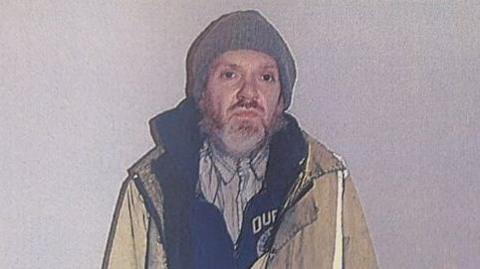 Roy Barclay, a bearded man with brown eyes, is in a yellow hi-vis jacket. He is also wearing a grey beanie, a striped shirt, and a dark hoodie with a logo or writing which is not fully visible behind his jacket.