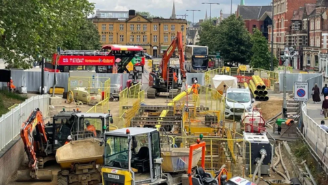 A cordoned-off section of the Botley Road ongoing works shows diggers and vans crowded around sections of road which have been dug up and are being supported by metal beams. In the background buses can be seen at bus stops and passengers can be seen walking along the roadworks cordon.