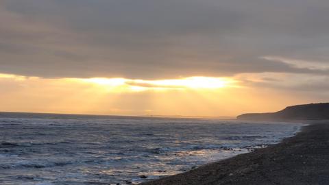 A general view looking out to the sea. In the foreground there is a narrow section of beach which is in the shade. There are thick clouds in the sky and sun rays are coming through, giving a shimmer to the small waves. On the horizon there is a cliff, also hit by the weak rays.