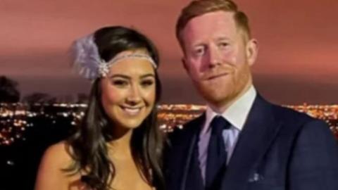 A woman and a man stand side by side. The woman has long dark hair and is wearing a hair band. The man has ginger hair and a ginger beard. He is wearing a dark suit with a white shirt