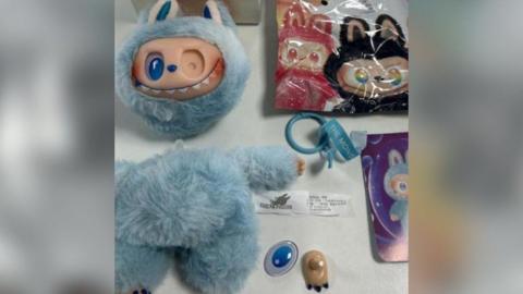 A fake Labubu doll - a gremlin style furry toy - with light blue fur and a face with many teeth sits on a white table. It is headless and missing eyes and feet and other features, demonstrating its danger to children. 