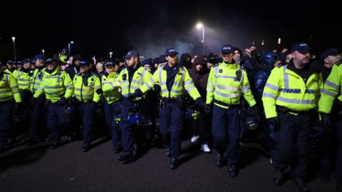 A line of more than a dozen police officers in high-visibility jackets and police hats. They are linking arms in front of a large crowd of football supporters, all of whom are wearing dark clothing. 