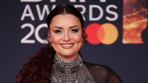 Shona McGarty smiles at the camera while on the red carpet at the Brit Awards. Her hair is tied back and she wears silver chunky earrings and a chain mail style vest over a see-through black blouse.