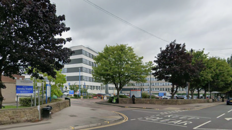 A black and white striped modern building in the background, with a row of trees and a road in the foreground. To the left is a blue and white sign which says "Barnsley Hospital".