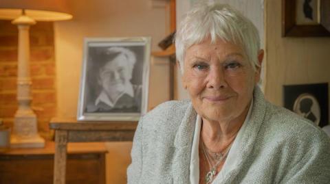 A woman with white hair and blue eyes smiling into the camera. She is wearing a grey blazer and silver necklaces. A black and white photograph of a man in a silver frame can be seen behind her.