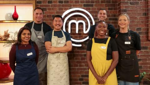 Three men and three women stand in front of a MasterChef sign, all wearing different coloured aprons
