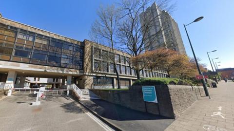 A street view image of Oldham Council's buildings which are a brown/grey and 1970s style with a flat roof. A small sign situated near the pavement reads Welcome to Oldham Council and there are red and white automatic barriers to stop cars getting in and out