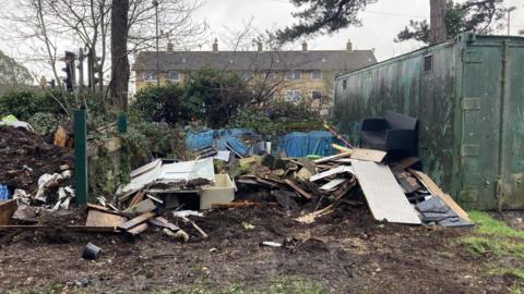 A large pile of waste, including wooden panels, planks and a black chair, are strewn across the ground. A large green shipping container can be seen next to the rubbish. 