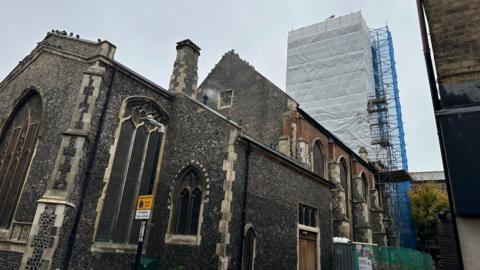 A black stone medieval church building with a tower in the background - scaffolded in white and blue.