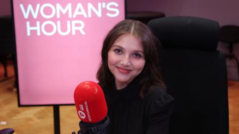 Mia McKenna-Bruce has long brown hair and wears black, and sits in the Woman's Hour studio