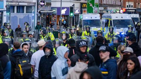 A large group of people standing near riot police, who are armed with shields and helmets. There are several police vans parked nearby some shops along a high street.