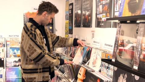 Gary Moore, manager of Assai Records in Dundee, sifts through stacks of vinyls displayed along a wall. He is wearing a checked brown cardigan and black t-shirt.