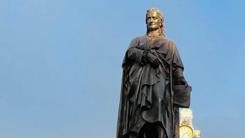 A close-up of a bronze statue of Sir Isaac Newton standing against a blue sky. It depicts him with long hair and wearing a coat, which seems to resemble robes. A clock tower can be seen in the background. 