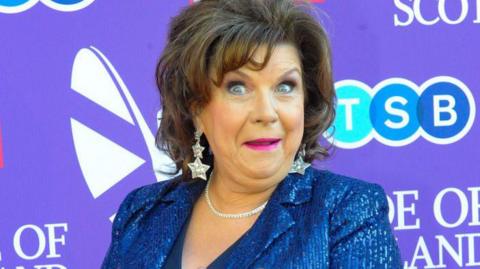 Elaine C Smith in sparkly blue jacket, pulling a face at the camera. She is standing in front of a purple background