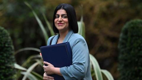 Shabana Mahmood, smiling while walking past plants, wearing a light blue jacket and holding a blue file.