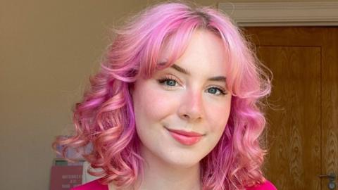 A woman with wavy pink hair in a pink top smiles at the camera for a selfie, taken indoors