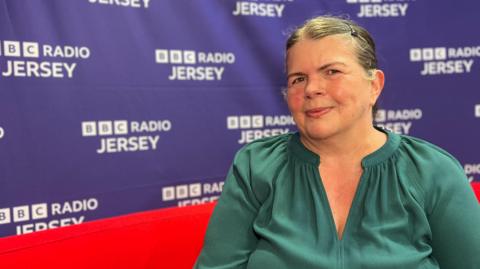 Sharon McLucas, who has her hair tied back and is in a green top, is sitting on red sofa with BBC branding behind her.