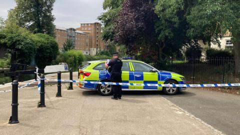 A police car is parked across a footpath near a river. A police officer is checking paperwork with his back to the camera. Blue and white police scenes of crime tape has been placed in front of him attached to a metal bollard to the left and iron fencing to the right.
A block of flats is visible in the background on the other side of the river.
