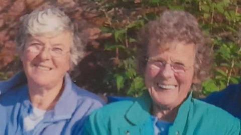 Maureen Bedford and Eileen Macleod smiling at the camera. They are photographed outside and both are wearing glasses and have short grey hair. 