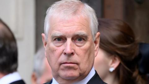 Andrew Mountbatten-Windsor in a black suit, white shirt and black tie looks directly at the camera while standing among other formally dressed people.