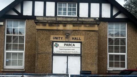 The exterior of a building, showing three windows with white window panes. The front is a mixture of brown stucco and at the top is white stucco with black beams. There is a sign that reads "Unity Hall 1927" and beneath it is a sign that reads "PARCA: Supporting people in need".