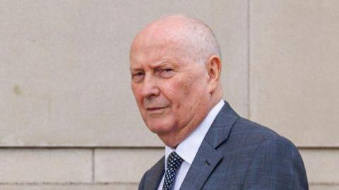 Cushnahan is wearing a navy blue tartan suit. He is wearing a white shirt and a black and white spotty tie. 